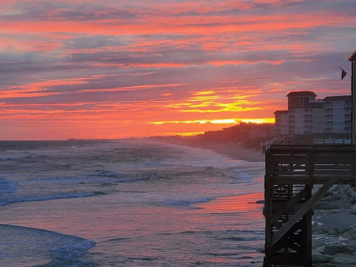 A wide view of the coastline at sunset, with the pier extending into the ocean, buildings along the shore, and soft waves reflecting the colors of the sky. The mood is peaceful and reflective, ideal for a sunset walk or sharing a special moment by the water.