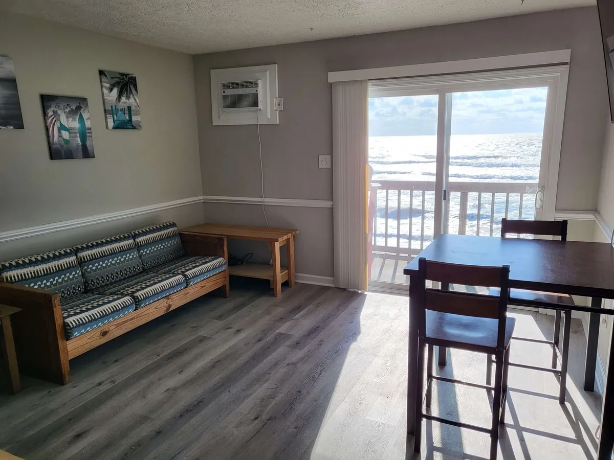 This image presents a cozy living area with a patterned sofa and a small dining table, all set near a large sliding glass door that opens to sweeping views of the beach and ocean. Sunlight pours into the space, creating a casual, inviting atmosphere ideal for relaxing, socializing, or enjoying meals with a view.