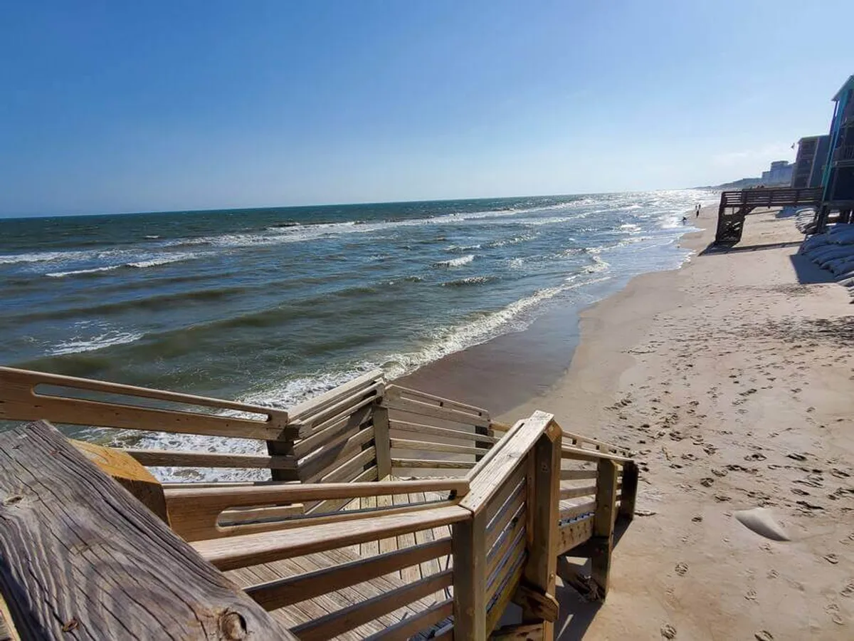 A wooden staircase leads down to a sandy beach, with ocean waves and distant buildings visible. The setting is serene and highlights seamless movement from the property to the beach.