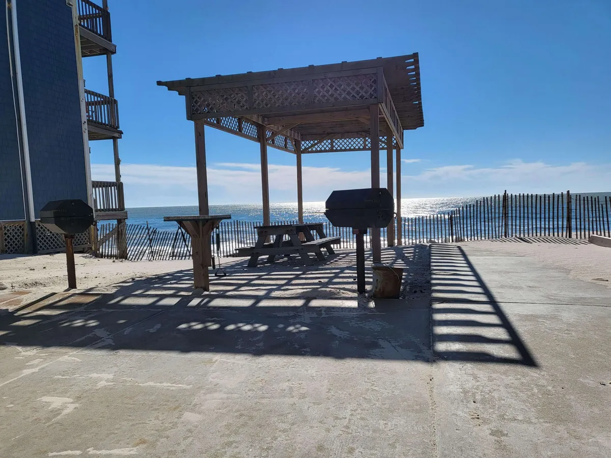 This image displays an inviting outdoor picnic area with a grill, picnic table, and sweeping ocean views in the background. The shadows cast by the gazebo add a relaxed coastal ambiance, suggesting a place where friends or solo travelers can gather, dine, or simply enjoy the scenery.