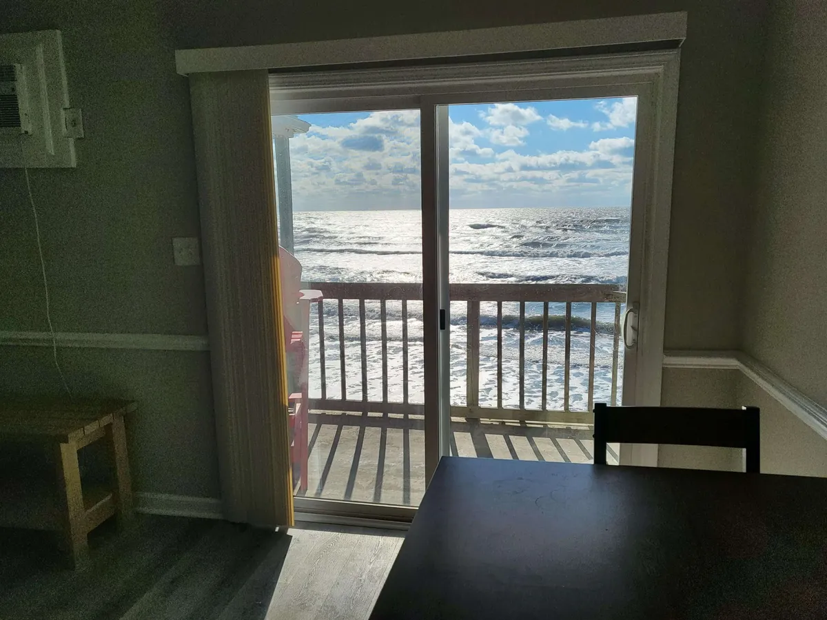 The photo displays a peaceful dining area next to a large sliding glass door, with a table set for enjoying meals or quiet moments while gazing out at the ocean. The view through the glass is mesmerizing, emphasizing the closeness to the beach and the opportunity to enjoy the ocean breeze from inside.