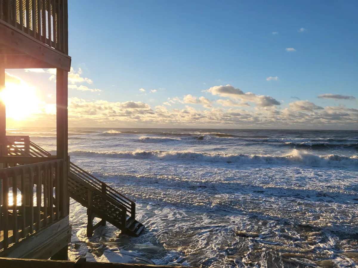 A tranquil scene showing a stunning sunset over the ocean, with waves gently crashing on the shore and a staircase leading down to the beach. The soft pastel hues of twilight create a romantic atmosphere perfect for couples enjoying an evening walk or a quiet moment together.