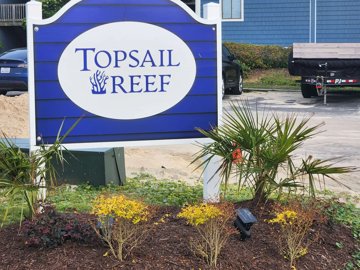 A tranquil exterior shot of the property entrance with the Topsail Reef sign, attractive landscaping, and surrounding vehicles. The setting is peaceful and welcoming, suggesting arrival and access to the area's natural beauty and community events.
