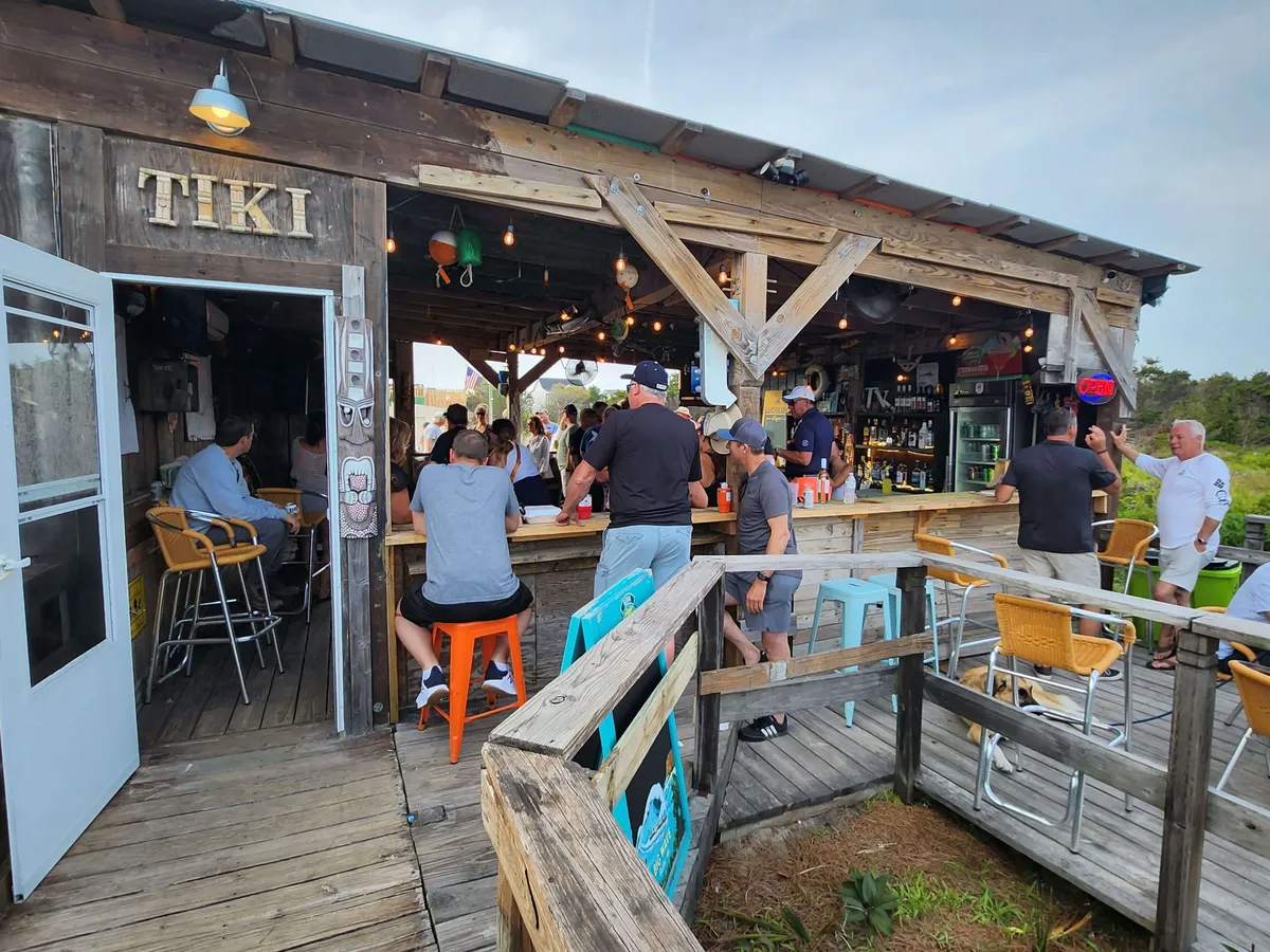 A vibrant outdoor Tiki bar scene featuring a prominent Tiki sign, bar area, outdoor seating, and groups of people socializing. The backdrop hints at sunset views and live entertainment, evoking the energy of local nightlife and community gatherings.