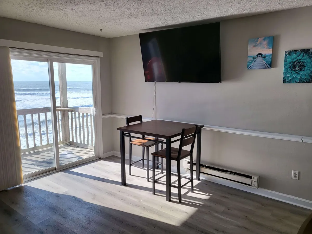 A serene interior shot featuring a dining table and chairs positioned by a sliding door that leads to the balcony, with a 65" Smart TV visible in the background. Beyond the glass, the ocean stretches out, offering breathtaking views from both the dining and relaxation areas.