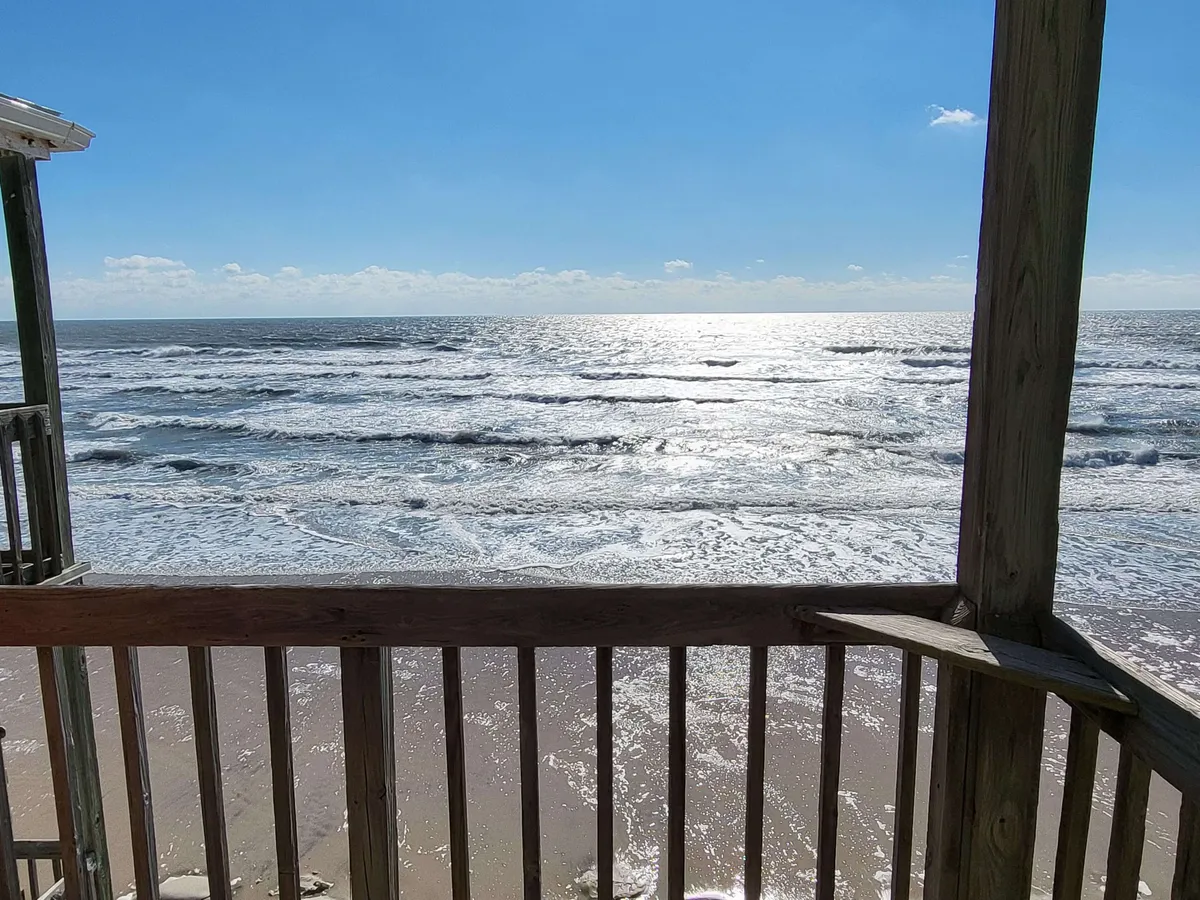 The image captures a serene view from the private balcony of the oceanfront condo, with gentle ocean waves rolling in, sunlight sparkling across the water, and a sturdy wooden railing framing the tranquil seascape. The mood is peaceful, inviting guests to relax, watch the sunrise or sunset, and enjoy the ocean breezes.