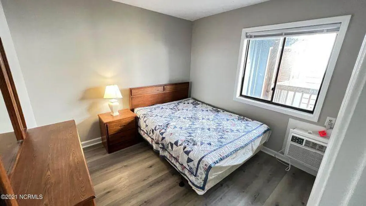 A bright, inviting bedroom with a plush bed, soft quilt, and a window letting in gentle natural light. A nightstand with a lamp sits beside the bed, creating a cozy and tranquil atmosphere ideal for relaxation and restful sleep.